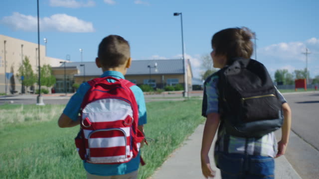 A Caucasian elementary-aged boy catches up to his friend as they both walk to school under a blue sky.