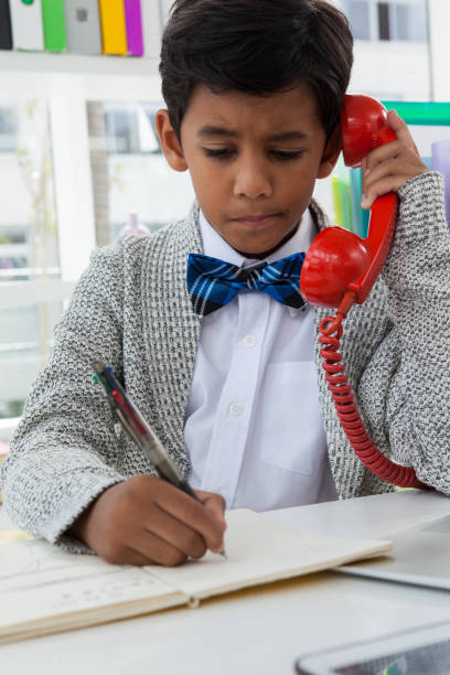 istock Boy imitating as businessman writing on book while using landline 825688390
