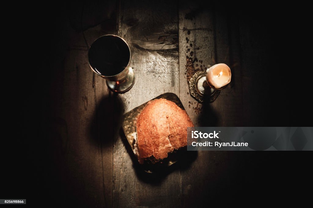 Bread and Wine Communion Sacraments A rustic wood table with old brass utensils holding broken bread and wine for the Catholic and Christian tradition of remembering the death and resurrection of Jesus Christ as he instructed at the last supper.  Dramatic lighting with candles. Communion Stock Photo