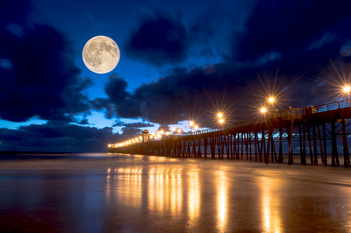 ocean pier at dusk with lights reflected in water and full moon in the cloudy sky