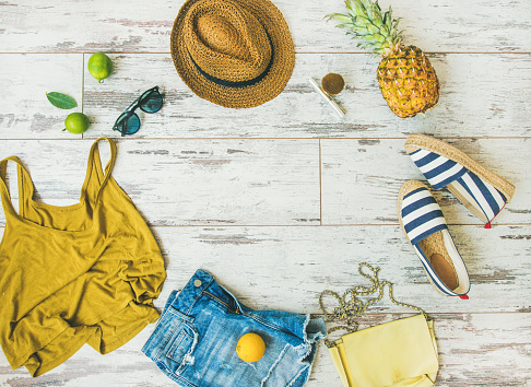 Colorful summer fashion outfit flat-lay. Denim shorts, straw sun hat, yellow top, espadrillas, leather bag, sunglasses, pineapple, limes and lemon over pastel parquet background, top view, copy space