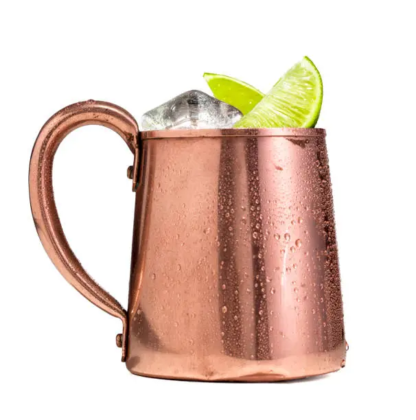 A Moscow mule drink in a copper cup on a white background.  Moscow mules are made with vodka, ginger beer, and lime and served over ice in a copper mug.