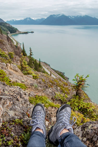 View of VIew of the Turnagain arm and Sewrard highs with Chugach mountain on the far background and female's feet in hiking boots on the foreground. Shot in the USA, Alaska, in summer. chugach mountains photos stock pictures, royalty-free photos & images