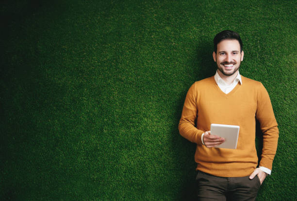 Attractive man standing over a green grass wall, holding a tablet