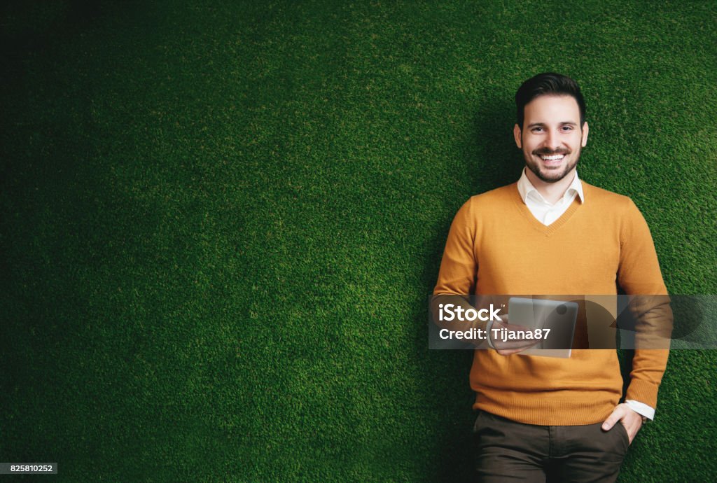 Attractive man standing over a green grass wall, holding a tablet Business Stock Photo