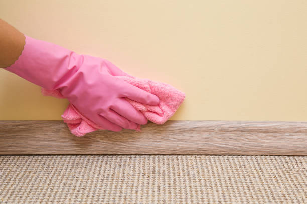 Hand in rubber protective glove with microfiber cloth cleaning baseboard on the floor from dust at the wall. Early spring cleaning or regular clean up. Maid cleans house. stock photo