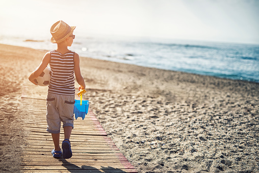 Summer vacations day morning. Little boy going to the beach to have fun. He is carrying a bucket and a ball to play with. The boy is looking at the sea. Lots of copy space.
