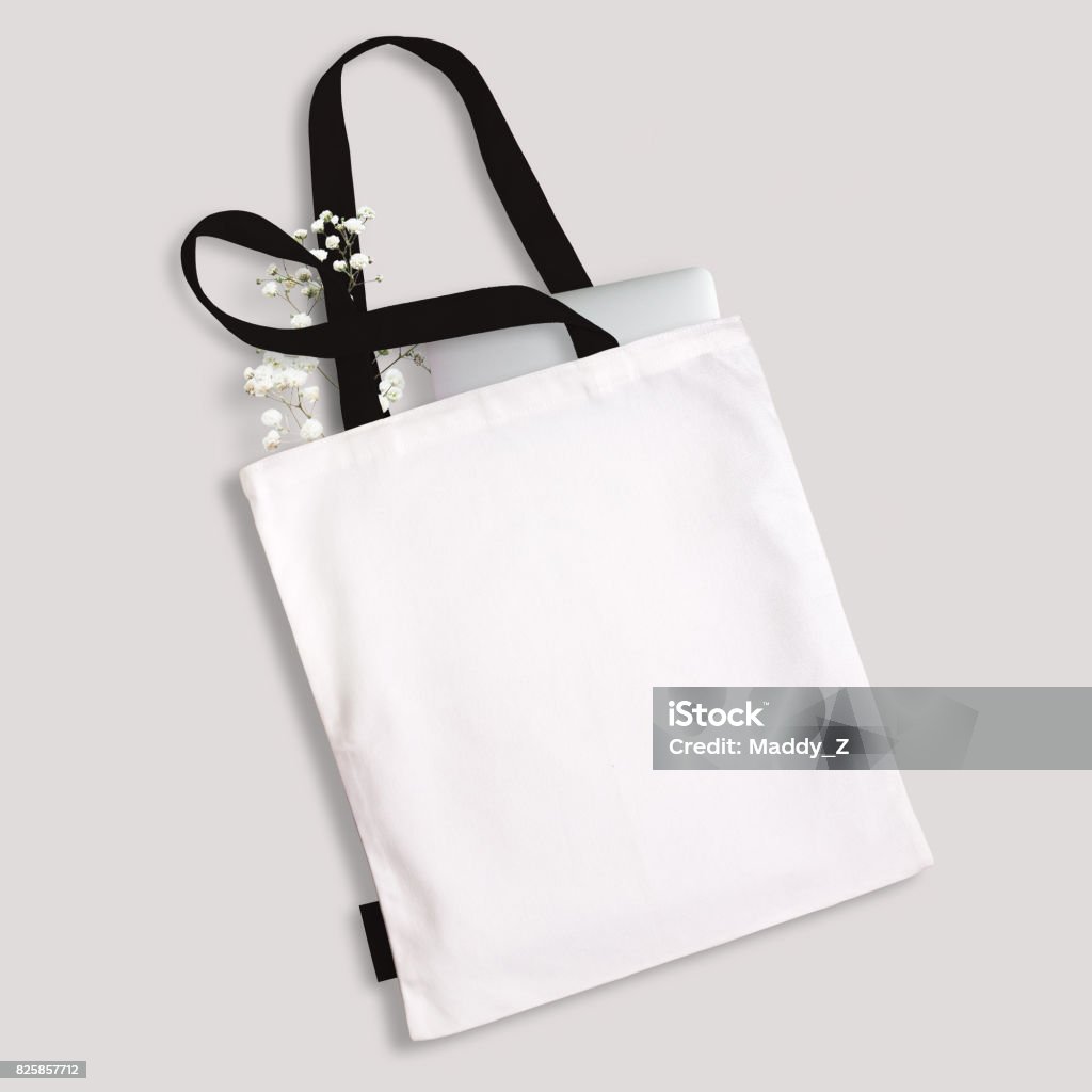 White blank cotton eco tote bag with black straps and little label, laptop and flowers inside. Design mockup White blank cotton eco tote bag with black straps, handles and little label, laptop and flowers inside. Design mockup. Bag Stock Photo