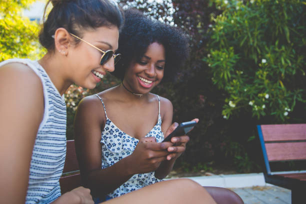 istock Young happy female friends sitting on bench using mobile 825870574