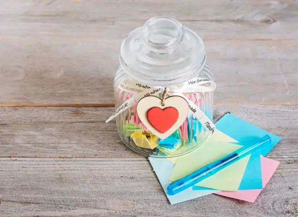 Empty glass jar with handmade wooden heart decoration and ribbon on a wooden plank with colorful pastel background.