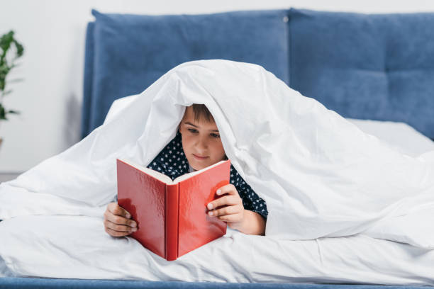 istock portrait of focused little boy reading book while lying under blanket in bed 825941602