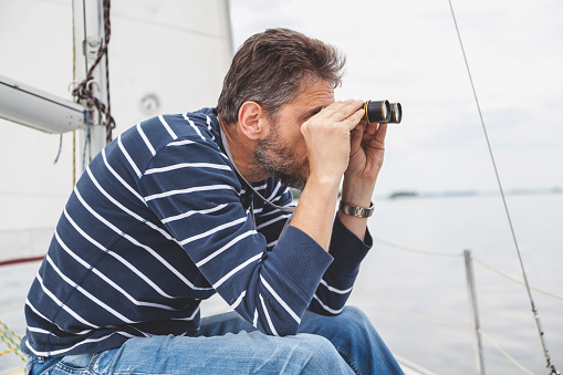 captain with a beard in a blue striped shirt sits on the deck of a sailing yacht and looks through close-up binoculars