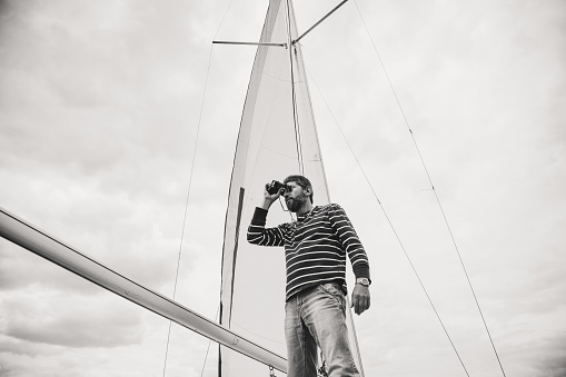 Monochrome image. Cute man with a beard with a striped shirt and jeans looking through binoculars sailing yacht on a background cloudy sky