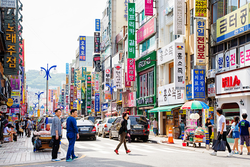 Busan South Korea Gwangbok-dong street scene daytime featuring the dense and colorful signs of boutiques and restaurants. Several people are seen on the sidewalk or crossing the street as as well as many cars.