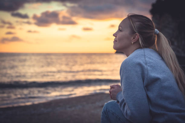 mujer sonriente solitaria pensativa mirando con esperanza en el horizonte durante el atardecer en la playa - i believe fotografías e imágenes de stock