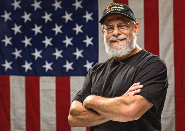 Authentic Vietnam Veteran with American Flag Bearded Vietnam veteran standing in front of an American Flag with arms crossed vet stock pictures, royalty-free photos & images
