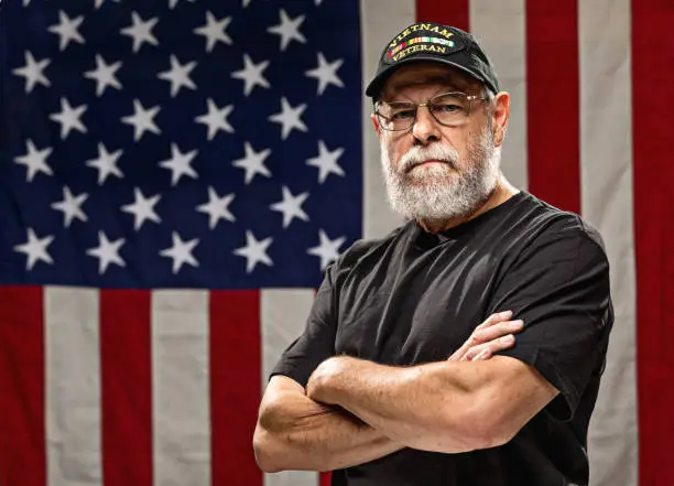 Bearded Vietnam veteran standing in front of an American Flag with arms crossed