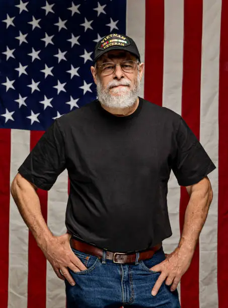 Bearded Vietnam veteran standing in front of an American Flag with arms crossed