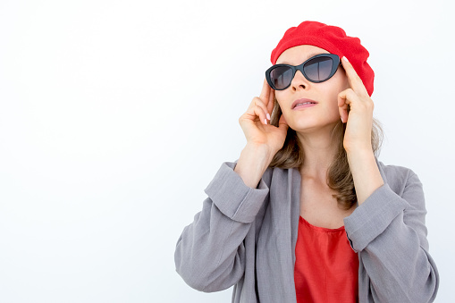 Enigmatic pensive woman in beret adjusting sunglasses and looking away. Strict French lady standing opposite white background. Elegance concept