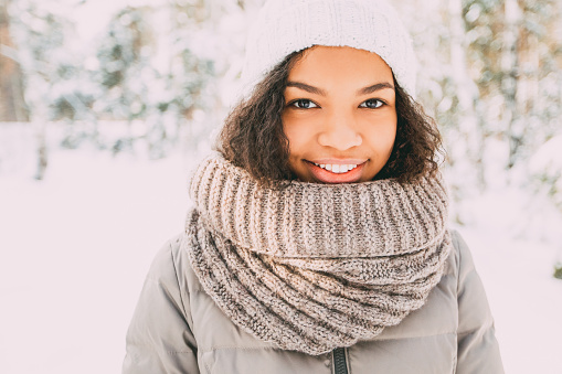 Portrait of happy young Latin American girl wearing white knit hat, scarf and jacket walking in winter, looking at camera and smiling