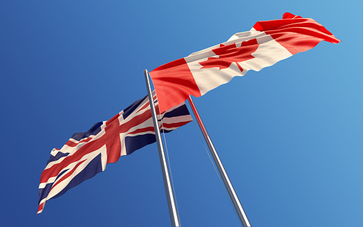 Canadian and British flags are waving with wind at opposite directions on a blue sky. Low angle view. Dispute and conflict concept. Horizontal composition with copy space. Great use for United Kingdom and Canada related concepts.