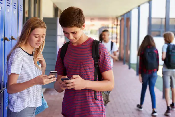 Photo of Two friends talking and using phones in school corridor