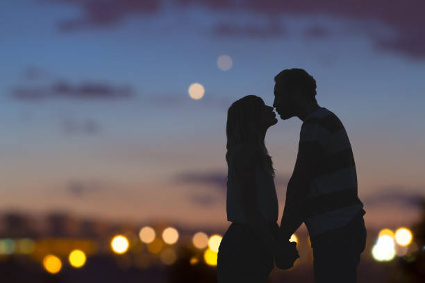 Photo of Silhouettes of a young couple kissing with city panorama in the background.