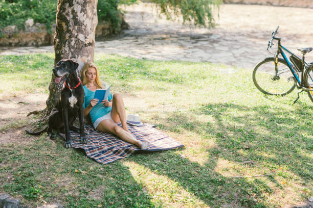 Mid adult woman enjoying the summer day with her dog, great dane, in the park. They are sitting on blanket and woman is reading book.