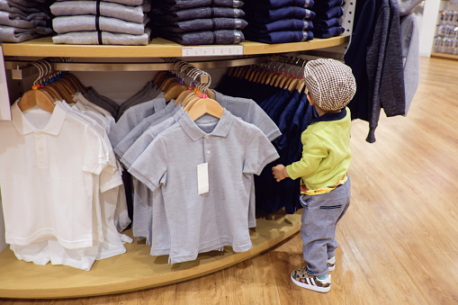 Cute little Asian 18 months / 1 year old toddler baby boy child choosing & buying kids clothes in garments shop, Seasonal sale, shopping. Kid's fashion. Modern children.