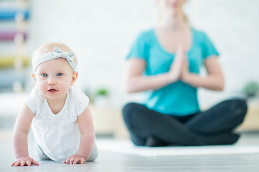 A mother is doing yoga in an indoor studio on a yoga mat. In this photo the baby girl is in the front of the frame while her mother is meditating in lotus position.