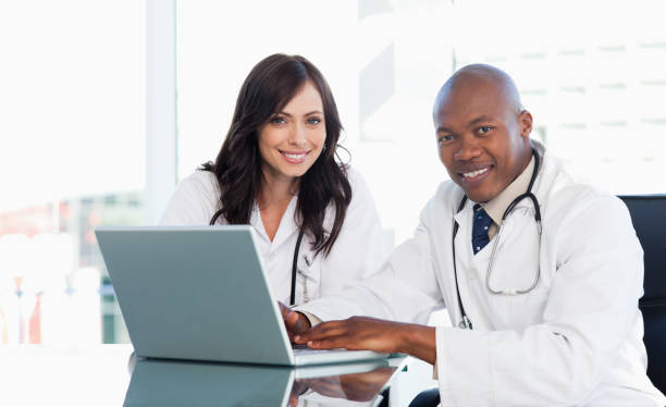 Smiling medical staff working in front of a laptop while sitting at the desk Medical staff working in front of a grey laptop while sitting at the desk in front of photos stock pictures, royalty-free photos & images