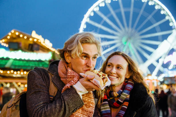 Eating At The Christmas Fair Happy couple are at the christmas fair and market. The man is eating a hotdog from one of the market stalls and his partner is laughing. hyde park london photos stock pictures, royalty-free photos & images