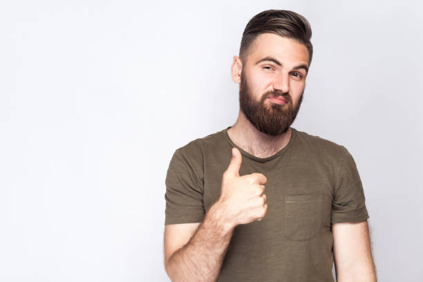 retrato de hombre con barba satisfecho con los pulgares para arriba y la camiseta verde oscuro contra el fondo gris claro. - confidence toothy smile thumbs up ok sign fotografías e imágenes de stock