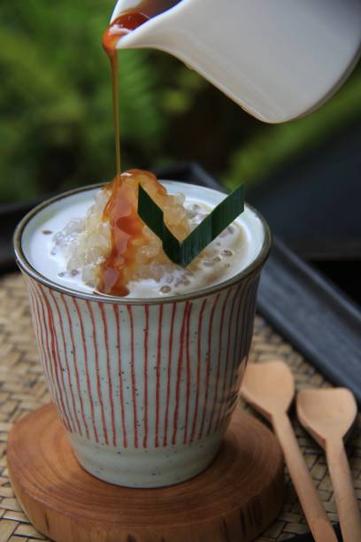 Sago Gula Melaka, a Traditional Malaysian Dessert of Sago Tapioca Pearls with Coconut Milk and Palm Sugar Sago Gula Melaka, a popular Malaysian dessert of sago tapioca pearls with coconut milk and palm sugar syrup. It is served in a tall ceramic cup with teakwood coaster. Here shown the sugar syrup is being poured onto the pearls. Two wooden teaspoons are placed next to the cup. A woven bamboo tray is used to serve the food, which took place outdoor in the terrace. johor photos stock pictures, royalty-free photos & images