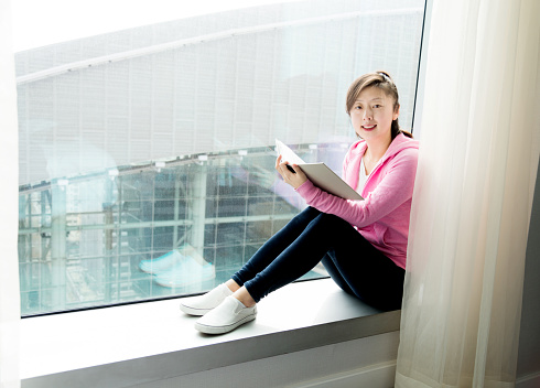 Young asian woman reading a book while sitting on window sill.