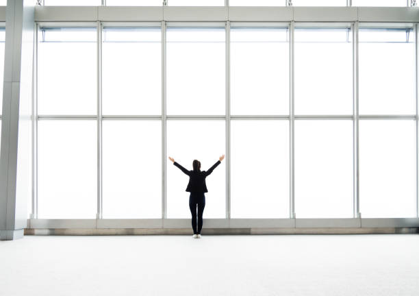Rear view of businesswoman standing in front of office window and arms outstretched.