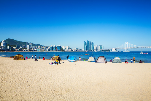 Gwangalli beach day scene in Busan, South Korea with the modern city in the background and many tents in the foreground on the sand. Many people are enjoying this Springtime day at the beach.