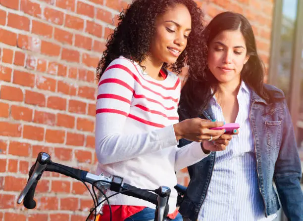 two young women friends standing near a brick wall with their bicycle looking at smart phone