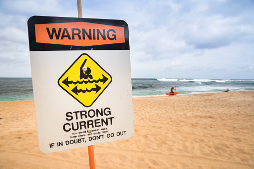 A sandy tropical beach with Rip Currents Sign and a breaking ocean wave in the background. Sign says: \