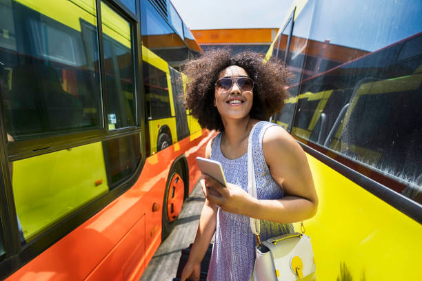 Cheerful tourist with smart phone and suit case walking through buses. Wears casual clothes.
