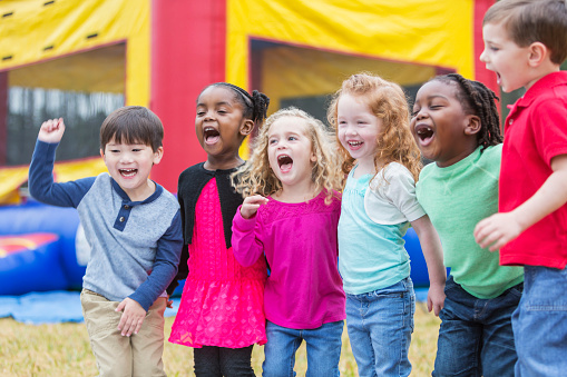 A group of six multi-ethic children, 3-4 years old, standing in the park together, laughing and shouting. A bouncy castle is in the background.