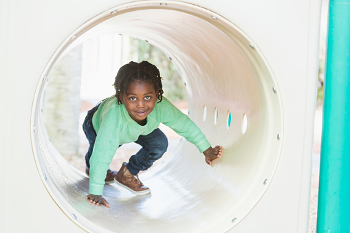 A little black 4 year old boy crawling through a tunnel on a playground, smiling at the camera.