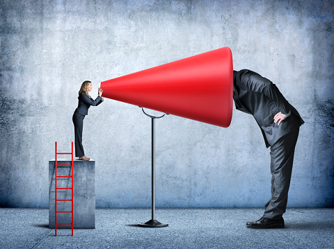 A businesswoman stands on a concrete pedestal as she shouts into a much large red megaphone in order to amplify her message.  A very large businessman sticks his head into the megaphone in order to hear better.