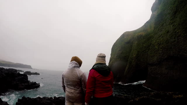 Back view of young tourists couple in raincoats standing on the shore of the sea in foggy day and enjoying the landscape