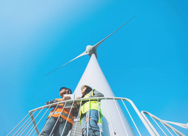 Maintenance engineers working on wind turbine in windfarm Low angle view of electrical engineer woman and technical businessman standing and working with digital tablet pc on high wind turbines at wind power energy generator station at offshore wind farm against blue clear sky with copy space area. XXXL Size Taken with Canon 5D MIV offshore wind farm stock pictures, royalty-free photos & images