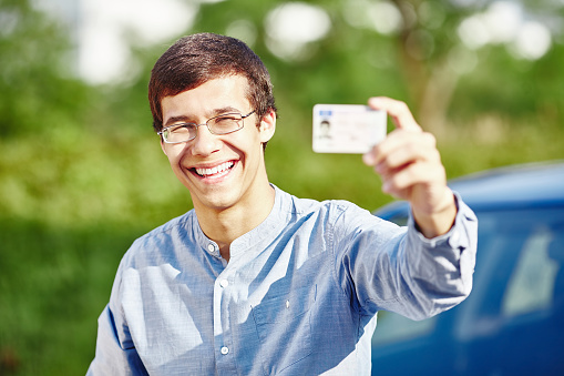 Close up portrait of young hispanic man wearing glasses and blue denim shirt holding out his driving license and smiling against car outdoors - new drivers concept