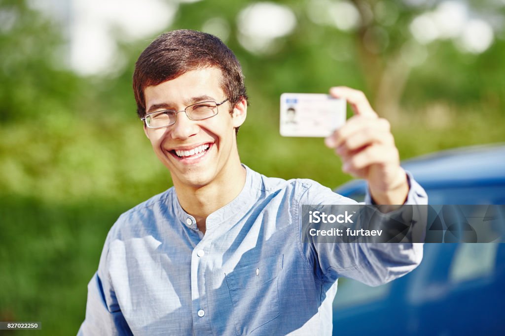 Guy with driving license Close up portrait of young hispanic man wearing glasses and blue denim shirt holding out his driving license and smiling against car outdoors - new drivers concept Driver's License Stock Photo