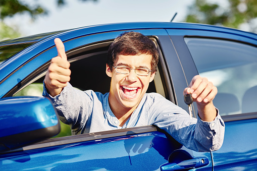 Close up of young happy hispanic man wearing glasses holding out car keys, showing thumb up hand gesture and laughing through car window - driving school and new drivers concept