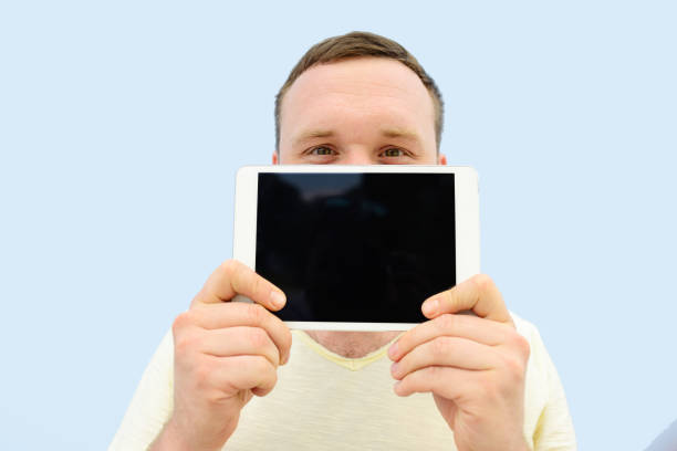 istock Handsome young man with a beard showing something on the tablet closing the face floor, isolated on a blue background, close-up for your text 827041190