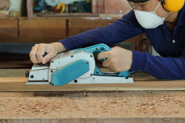 istock Carpenter using electric planer with wooden plank in carpentry workshop. He is wearing safety equipment 827067292